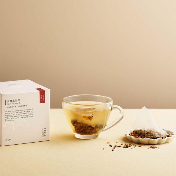 女性在室內自然光下手持玻璃杯飲用黑豆茶茶包,茶色溫潤清透,後方擺放產品包裝,呈現放鬆的日常飲茶情境
